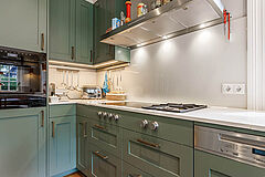 Landhausküche in Salbeigrün, Hamburg Altona | maßgefertigt von der Möbeltischlerei woodendesign – Küchenzeile mit Gaggenau Kochfeld, Rückwand aus Glas und Keramikarbeitsplatte