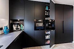Geöffneter Hochschrank mit Stauraum und Auszügen neben Backofen und Kaffeemaschine in der mattschwarzen Front.