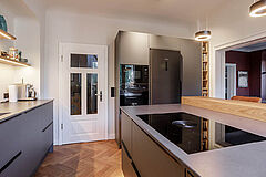 Kücheninsel mit BORA Induktionsfeld und Eiche-Sitztheke – Blick Richtung Hochschrankwand und Tür, hölzerner Fischgrätboden sichtbar.