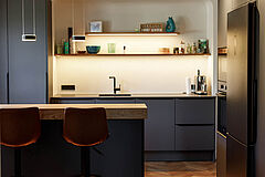 Einbauküche mit Sitztheke in Hamburg-Bergedorf – Blick auf Spülenzeile mit offenen, beleuchteten Regalen und die Insel mit Eiche-Theke im Vordergrund.