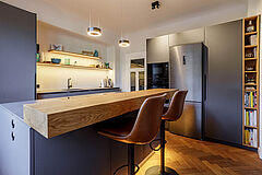 Kücheninsel mit Eiche-Sitztheke und zwei Barstühlen – Blickrichtung zu den hohen Schränken und der Spülenzeile.