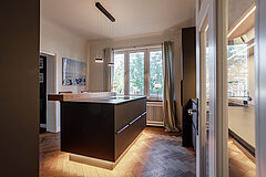 Einbauküche mit Sitztheke – seitlicher Blick auf die Insel mit Sockelbeleuchtung und Eiche-Theke, Fensterfront im Hintergrund.