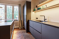 Spülenzeile der Einbauküche – dunkle Unterschränke, Armatur und offene Regale mit warmem Lichtband, Fenster und Vorhänge daneben.