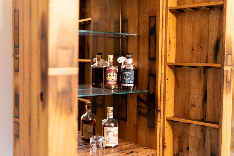 Innenansicht eines Altholz-Schranks als Hausbar: Glasböden mit Flaschen und Gläsern, seitliche Holzleisten, warmes Licht, ruhige Linien. Aktuelles – Trends – News im November 2025 | Möbeltischlerei woodendesign | Tischlerei Möbeltischler Möbelmanufaktur Hamburg