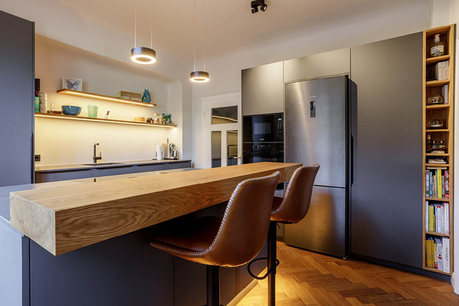 Küche mit offener Regalzeile und beleuchteten Wandborden; große Eichen-Inselplatte, seitliches Bücherregal, wohnliche Verbindung der Zonen. Aktuelles – Trends – News im November 2025 | Möbeltischlerei woodendesign | Tischlerei Möbeltischler Möbelmanufaktur Hamburg