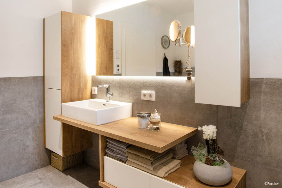 Bad mit weißem Waschtisch, Eichen-Ablage und Hochschrank; warmes LED-Licht, offene Handtuchablage, ruhiges Frontbild. Holz im Badezimmer | Aktuelles – Trends – News im Dezember 2025 | Möbeltischlerei Tischlerei Möbeltischler Möbelmanufaktur Hamburg