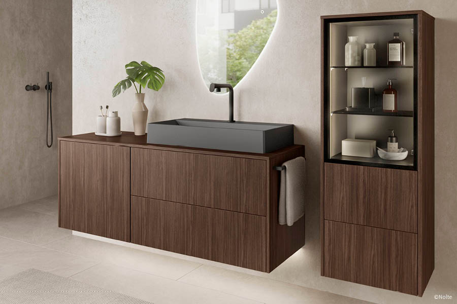 Dunkles Badmöbel-Ensemble: breiter Waschtischunterschrank in Nussbaum-Optik mit schwarzem Aufsatzwaschbecken, daneben beleuchtete Vitrinen-Säule. Holz im Badezimmer | Aktuelles – Trends – News im Dezember 2025 | Möbeltischlerei Tischlerei Möbeltischler Möbelmanufaktur Hamburg