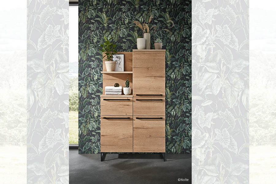Hochschrank in heller Eiche vor dunkler Blättertapete; offene Fächer und Türen, schwarze Griffleisten, wohnlicher Akzent. Holz im Badezimmer | Aktuelles – Trends – News im Dezember 2025 | Möbeltischlerei Tischlerei Möbeltischler Möbelmanufaktur Hamburg