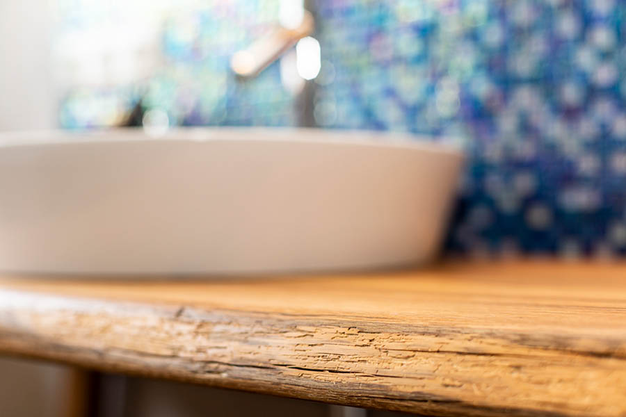 Detail Eichen-Waschtischplatte mit rustikaler Kante; unscharfes rundes Aufsatzwaschbecken davor, Mosaikfliesen im Hintergrund. Holz im Badezimmer | Aktuelles – Trends – News im Dezember 2025 | Möbeltischlerei Tischlerei Möbeltischler Möbelmanufaktur Hamburg