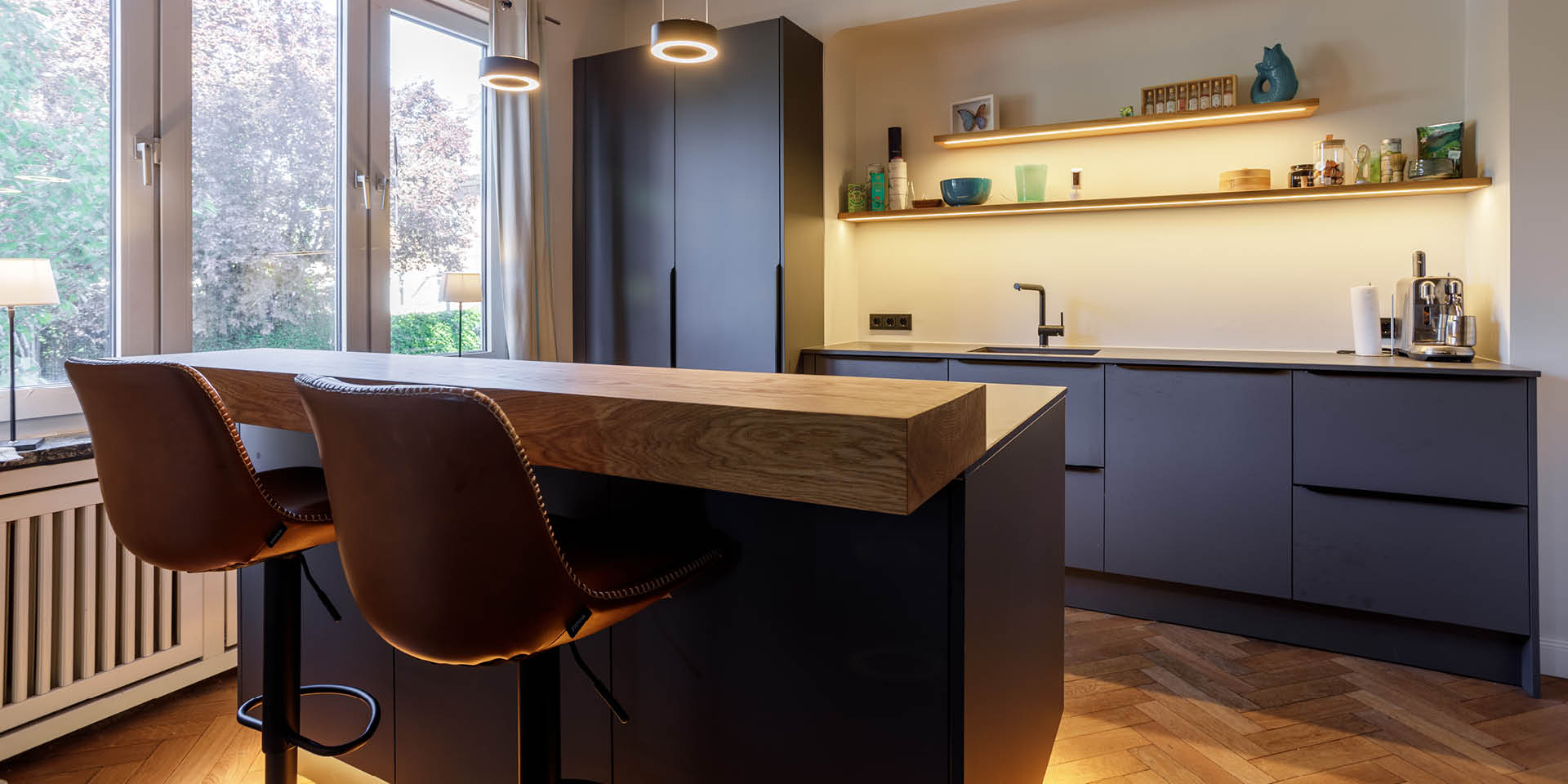 Einbauküche mit Sitztheke in Hamburg-Bergedorf – dunkle Fronten, Kücheninsel mit Eiche-Theke und Sitzplätzen, offene Regale mit Licht.